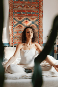 Woman sits cross‑legged on a bed with eyes closed, meditating, wearing white clothing in a sunlit room with a colorful textile wall hanging behind her.