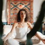 Woman meditating cross-legged on a white mat, eyes closed, in a room with a patterned wall hanging behind her.
