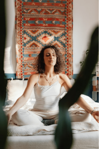 Woman meditating cross-legged on a bed, eyes closed, wearing a white top, in a room with a decorative textile tapestry behind her.