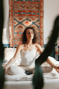 Woman meditating cross-legged on a white mat, eyes closed, in a room with a patterned wall hanging behind her.