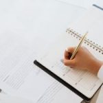 Person writes in a spiral notebook on a desk with scattered papers and a laptop nearby.