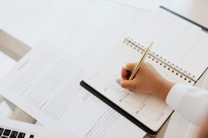 Close-up of a hand writing in a spiral-bound planner on a desk, with documents and a laptop nearby.