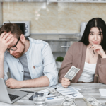 Stressed man with glasses rests his head on his hand next to a laptop; a woman sits with cash on a kitchen counter.
