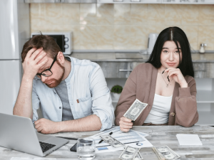 A man sits at a desk with his head in his hand, looking stressed, while a woman beside him holds a dollar bill and smiles slightly; money and documents are spread on the table in a kitchen-like setting.