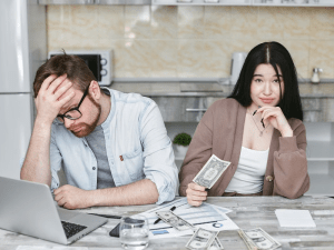 Stressed man with glasses rests his head on his hand next to a laptop; a woman sits with cash on a kitchen counter.